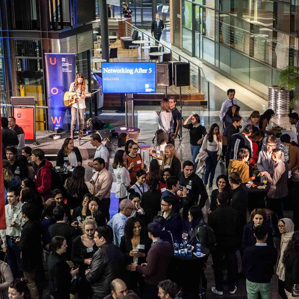 A large crowd of people mingling and conversing in front of a stage. On the stage, a musician plays a guitar. The backdrop features a blue screen displaying the words 